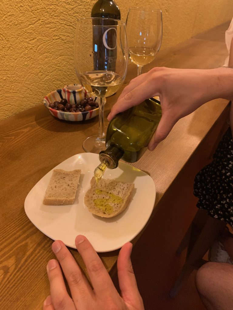 Manos vertiendo aceite de oliva virgen extra ecológico a pan casero en un plato blanco, con una botella de vino y dos copas de vino blanco al fondo, reflejando una experiencia gastronómica auténtica en Cortijo El Cura